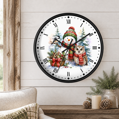 A round wall clock with a black frame and a festive winter illustration featuring a snowman in a red knit hat and scarf, an owl in a red scarf, Christmas trees, and ornaments. The clock is mounted on a light wood-paneled wall above a wooden table with a candle, pinecones, and a small plant, with a beige armchair partially visible in the foreground.