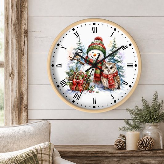 A round wall clock with a light wood frame and a festive winter illustration featuring a snowman, owl, evergreen trees, and a basket of ornaments. The clock is mounted on a white wood-paneled wall above a rustic wooden table with a candle, pinecones, and a small plant, with a beige armchair partially visible on the left.