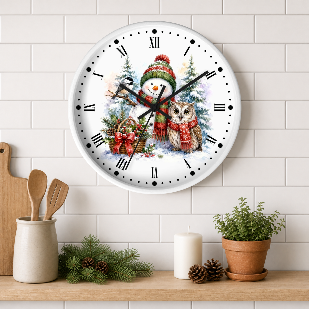 A round wall clock with a festive winter illustration featuring a snowman in a red knit hat and scarf next to an owl, mounted on a white subway tile wall above a wooden shelf with kitchen decor.