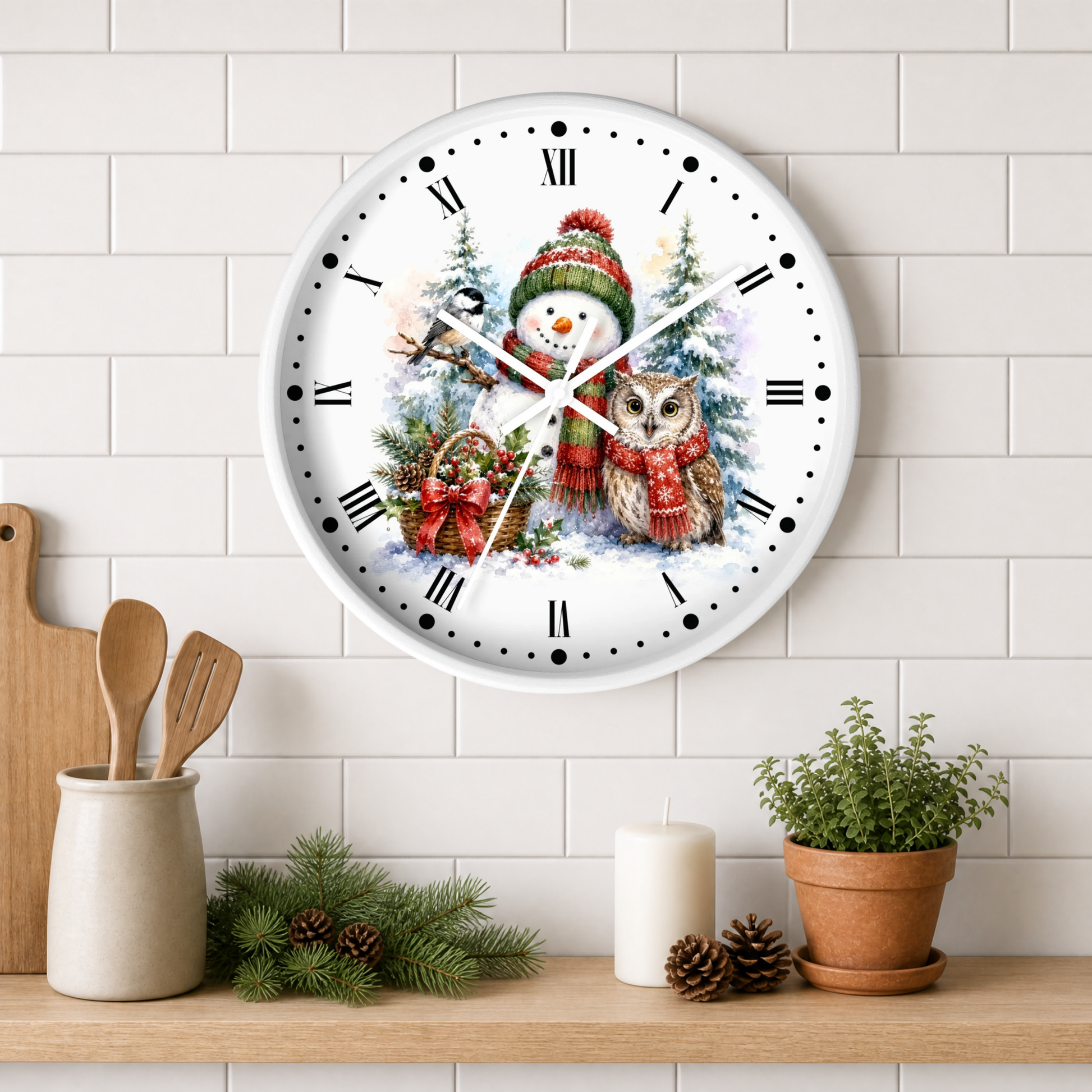 A round white wall clock with a festive winter illustration of a snowman and an owl, mounted on a white subway tile wall above a wooden shelf with kitchen decor and a potted plant.