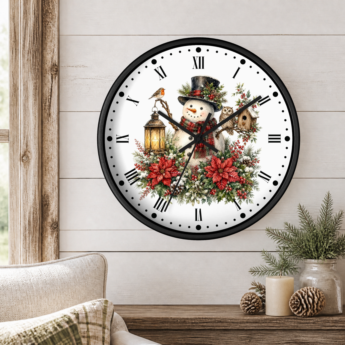 A round wall clock with a black frame and a white face, decorated with a festive Christmas design featuring a snowman, red poinsettias, green foliage, a bird, an owl, a lantern, and a birdhouse. The clock is mounted on a light wood-paneled wall above a wooden table with a candle and pinecones.