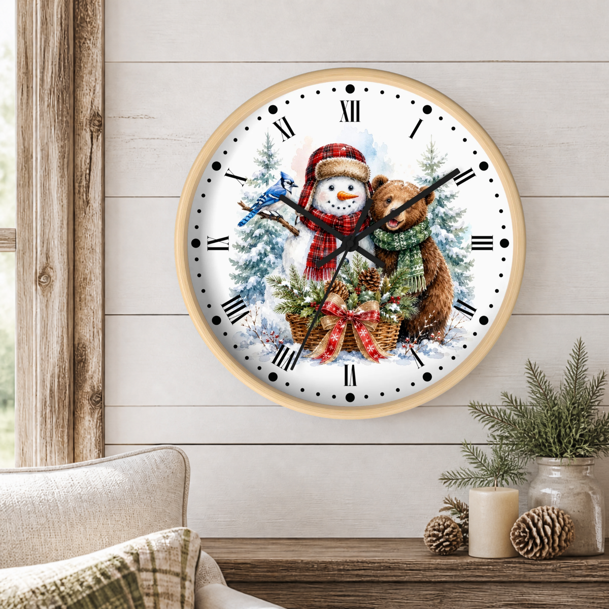 A round wall clock with a light wood frame hangs on a white shiplap wall above a wooden table. The clock face features a winter holiday illustration with a snowman in a red scarf, a brown bear in a green scarf, pine trees, and a bluebird.