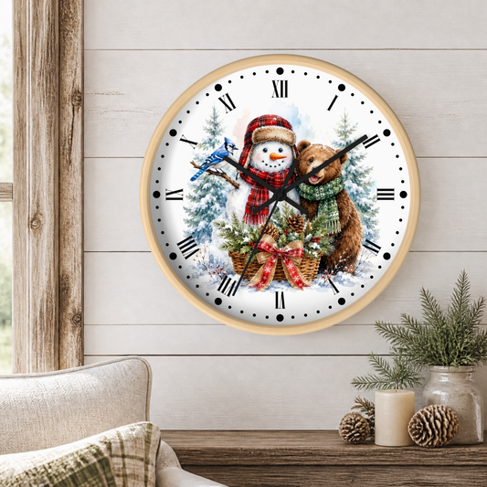 A round wall clock with a light wood frame hangs on a white shiplap wall above a wooden table. The clock face features a winter holiday illustration with a snowman in a red scarf, a brown bear in a green scarf, pine trees, and a bluebird.