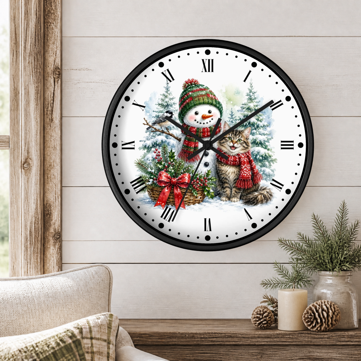 A round wall clock with a black frame and a festive winter illustration featuring a snowman and a cat in red scarves, displayed on a light wood paneled wall above a wooden table with candles and pinecones.