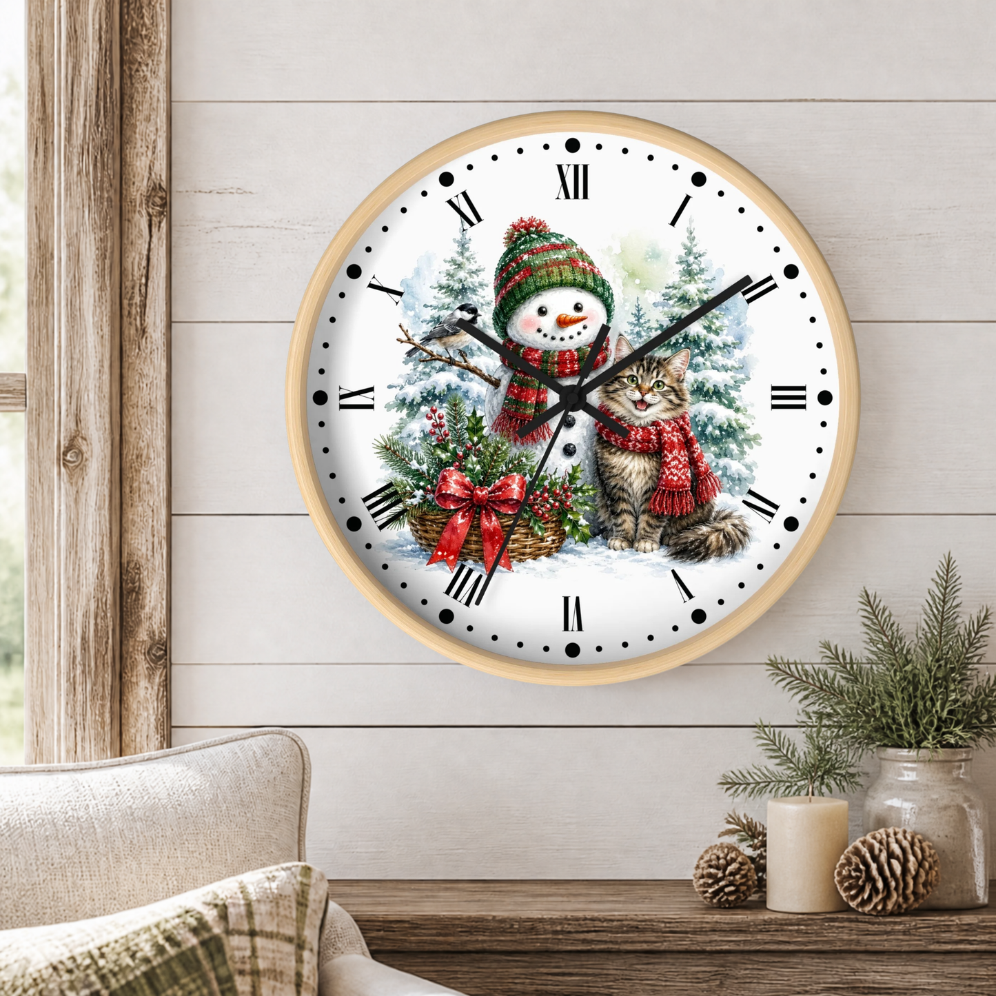 A round wall clock with a light wood frame hangs on a white shiplap wall above a wooden table. The clock face features a winter illustration of a snowman wearing a red and green knit hat and scarf next to a gray tabby cat in a red knit outfit.