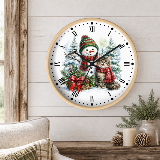 A round wall clock with a light wood frame hangs on a white shiplap wall above a wooden table. The clock face features a winter illustration of a snowman wearing a red and green knit hat and scarf next to a gray tabby cat in a red knit outfit.