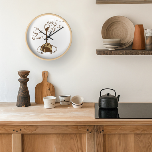 A round wall clock with a light wood frame hangs on a cream wall above a wooden kitchen counter. The clock face is white with a black line drawing of a hand holding a spoon over a pastry and the text 'The Joy of Autumn.
