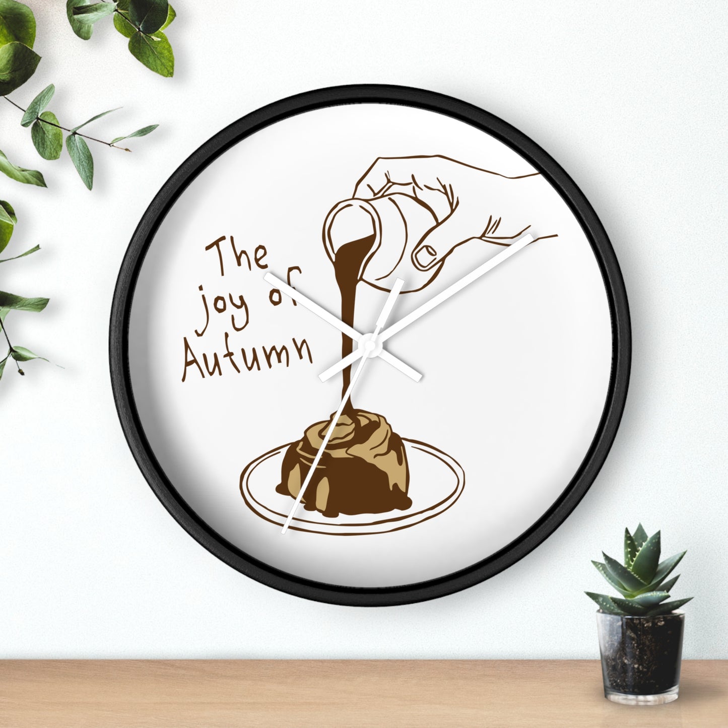 A round wall clock with a black frame and a white face, featuring a brown illustration of a hand pouring chocolate sauce over a dessert with the text 'The joy of Autumn'. The clock is mounted on a white wall above a light wood surface, with decorative green leaves and a small succulent plant nearby.