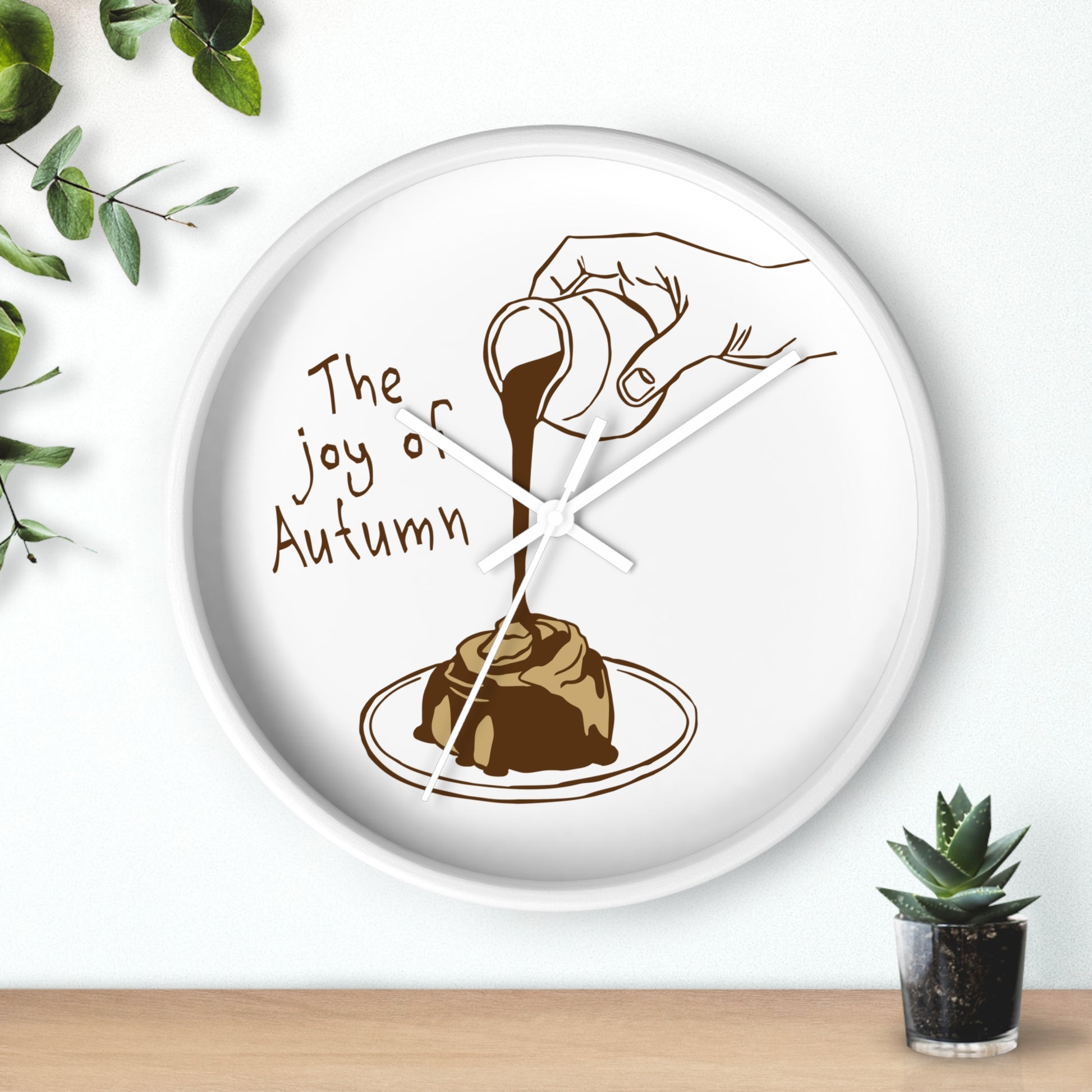 A round white wall clock with a brown illustration of a hand pouring chocolate pudding over a plate, with the text 'The joy of Autumn' on a white background. The clock is styled on a wooden surface with green leaves and a small succulent plant.