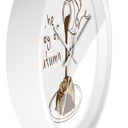 A white round wall clock with a minimalist autumn-themed design. The clock face features a brown illustration of a hand holding a teacup pouring tea into a rolling pastry on a plate, accompanied by the cursive text 'the joy of autumn' in brown.