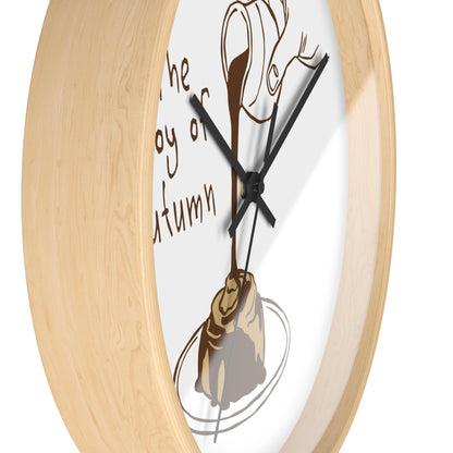 Close-up of a round wall clock with a light natural wood frame and a white face featuring a brown illustration of a pastry and dripping chocolate, with partial text reading 'The joy of autumn'.