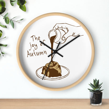A round wall clock with a light wood frame and a white face, featuring a brown illustration of a hand pouring syrup over a pastry with the text 'The joy of Autumn'. The clock is mounted on a white wall above a wooden surface, with green plants in the corners.