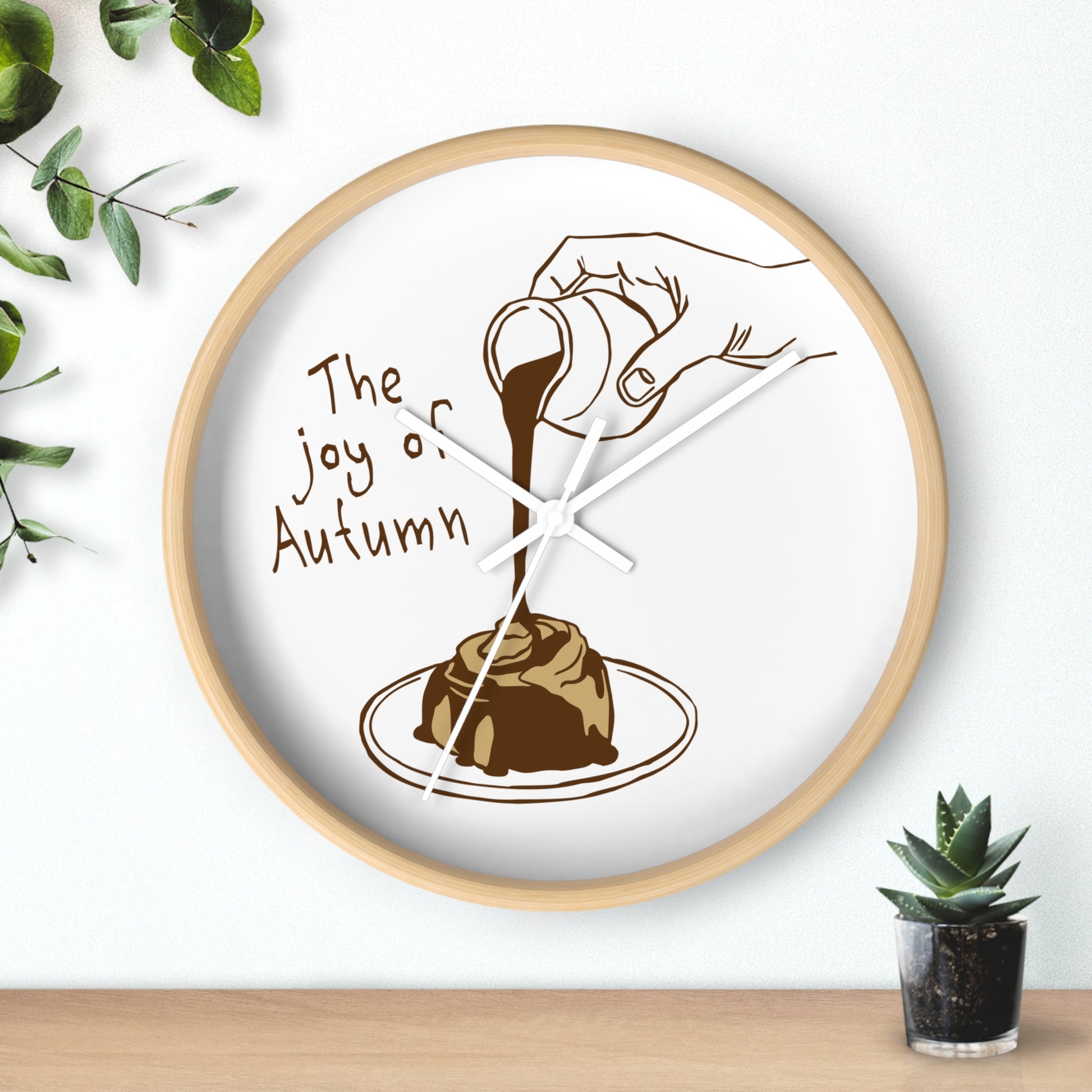 Round wall clock with a light wood frame and a white face, featuring a brown illustration of a hand pouring chocolate sauce over a dessert with the text 'The joy of Autumn'. The clock is styled on a light wood surface against a white wall with decorative green leaves and a small succulent.