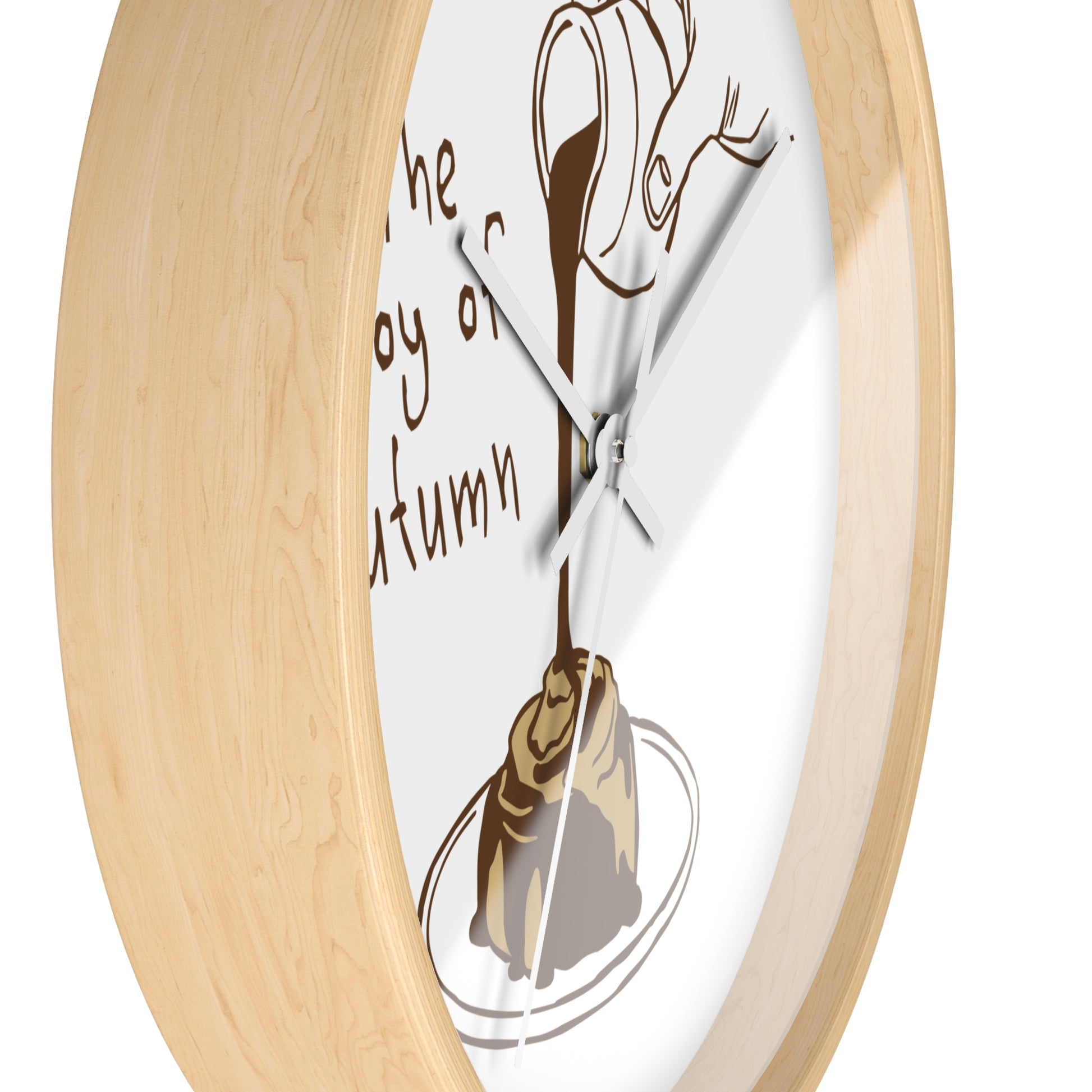 A close-up of a round wall clock with a light natural wood frame and a white dial. The dial features a brown illustration of a dripping coffee cup pouring into a pastry on a plate, with partial brown script text on the left.