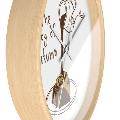 A close-up of a round wall clock with a light natural wood frame and a white dial. The dial features a brown illustration of a dripping coffee cup pouring into a pastry on a plate, with partial brown script text on the left.