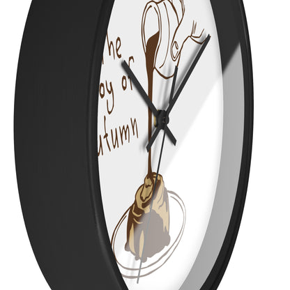 A close-up of a round wall clock with a black frame and a white face. The clock face features a brown illustration of a coffee cup pouring into a coffee bean on a plate, with partial brown text reading 'the joy of autumn'.