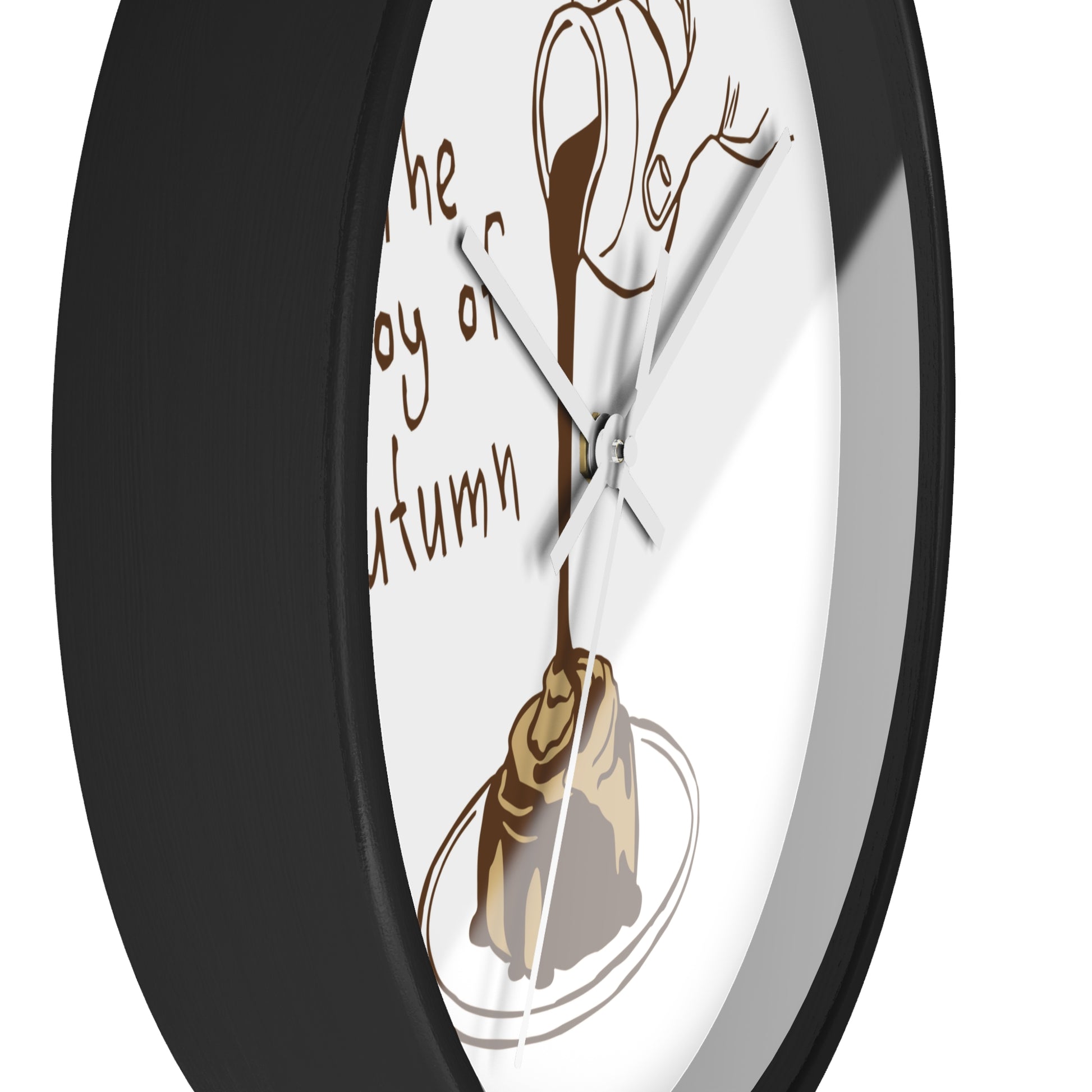 A close-up of a round wall clock with a black frame and a white face. The dial features a brown illustration of a cinnamon roll on a plate with dripping frosting and partial cursive text reading 'the joy of autumn.