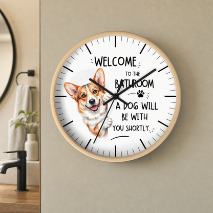 A round wall clock with a light wood frame hangs on a beige wall in a bathroom setting. The clock face is white with black hands and hour markers, featuring a watercolor illustration of a corgi and the text 'WELCOME TO THE BATHROOM A DOG WILL BE WITH YOU SHORTLY'.