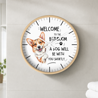 A round wall clock with a light wood frame and white face, featuring a Corgi illustration and the text 'WELCOME TO THE BATHROOM A DOG WILL BE WITH YOU SHORTLY' mounted on a light gray tiled wall.