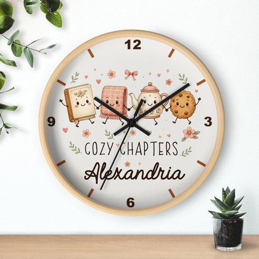 A round wooden wall clock with a white face, featuring cute illustrated characters and the text 'COZY CHAPTERS Alexandria'. The clock is shown hanging on a white wall above a wooden shelf, with small green plants in the corners.