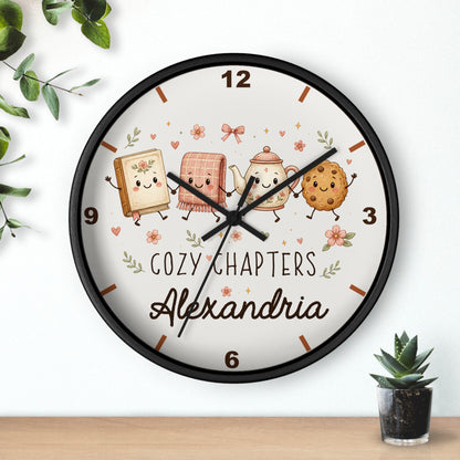 A round wall clock with a black frame and a whimsical cozy book-themed design. The clock face is cream-colored with cute illustrated characters (a book, a cozy blanket, a teapot, and a cookie) and the text 'COZY CHAPTERS Alexandria'.