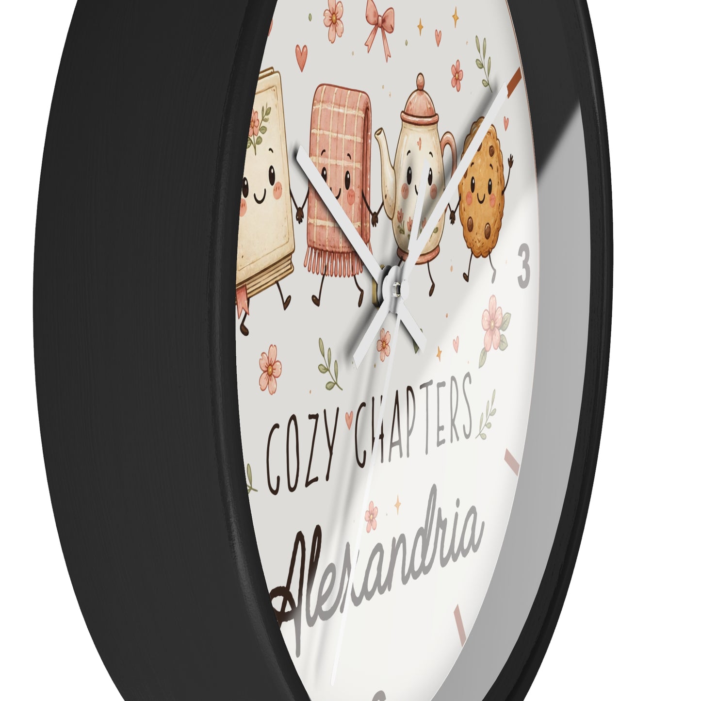A round wall clock with a matte black frame and a white face featuring cute illustrated book, tea towel, teapot, and cookie characters, plus the text 'COZY CHAPTERS' and 'Alexandria'.