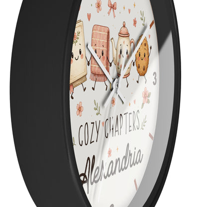 A round wall clock with a matte black frame and a white face featuring cute illustrated book, tea towel, teapot, and cookie characters, plus the text 'COZY CHAPTERS' and 'Alexandria'.