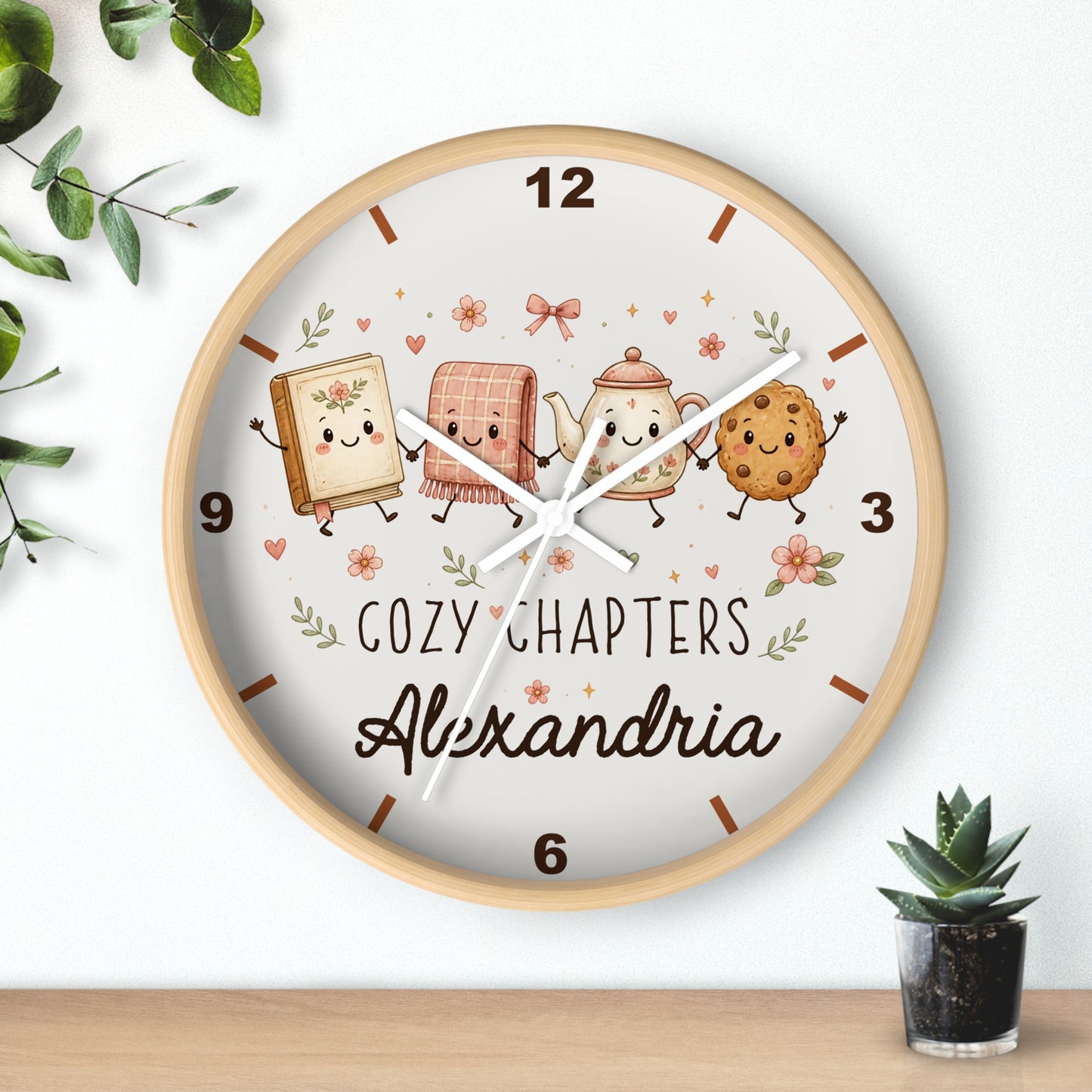 A round wooden-framed wall clock with a white face, featuring cute illustrated characters and the text 'COZY CHAPTERS' and 'Alexandria'. The clock is shown hanging on a white wall above a wooden surface, with small green plants in the corners.