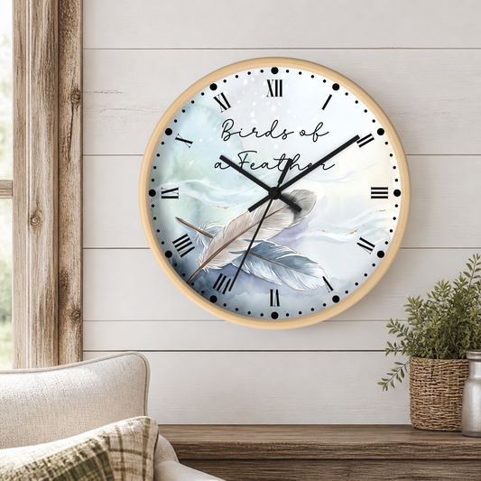 A round wall clock with a light wood frame hangs on a white shiplap wall above a wooden table. The clock face features a soft blue and white illustration of a pair of  feathers, black Roman numerals, and the words 'Birds of a Feather' in script and cursive.