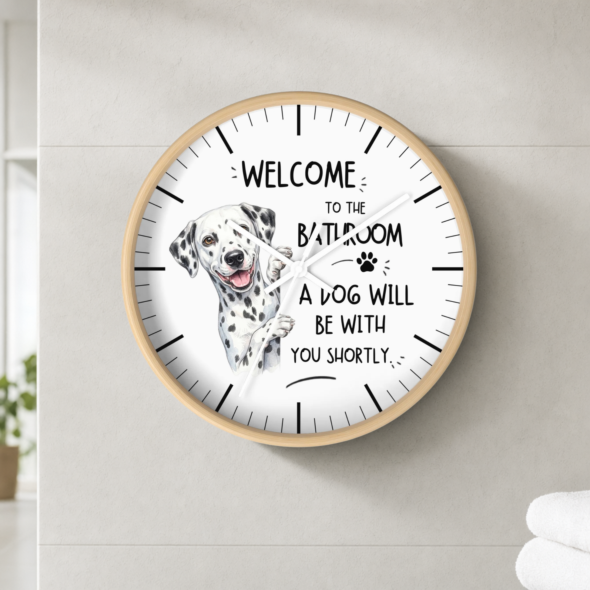 A round wall clock with a light wood frame and a white face, mounted on a light gray tiled wall. The clock features a Dalmatian illustration and the humorous text 'WELCOME TO THE BATHROOM A DOG WILL BE WITH YOU SHORTLY'.