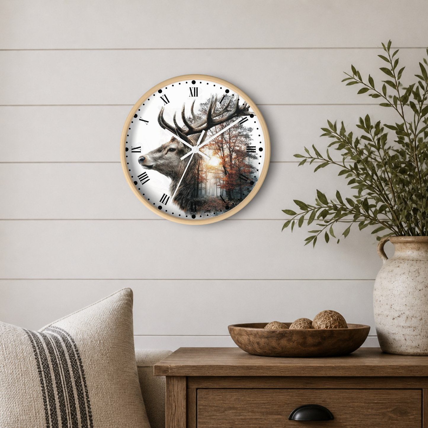 Round wall clock with a light wood frame and a printed deer and forest scene on the face, mounted on a white shiplap wall above a wooden side table with a vase and bowl.