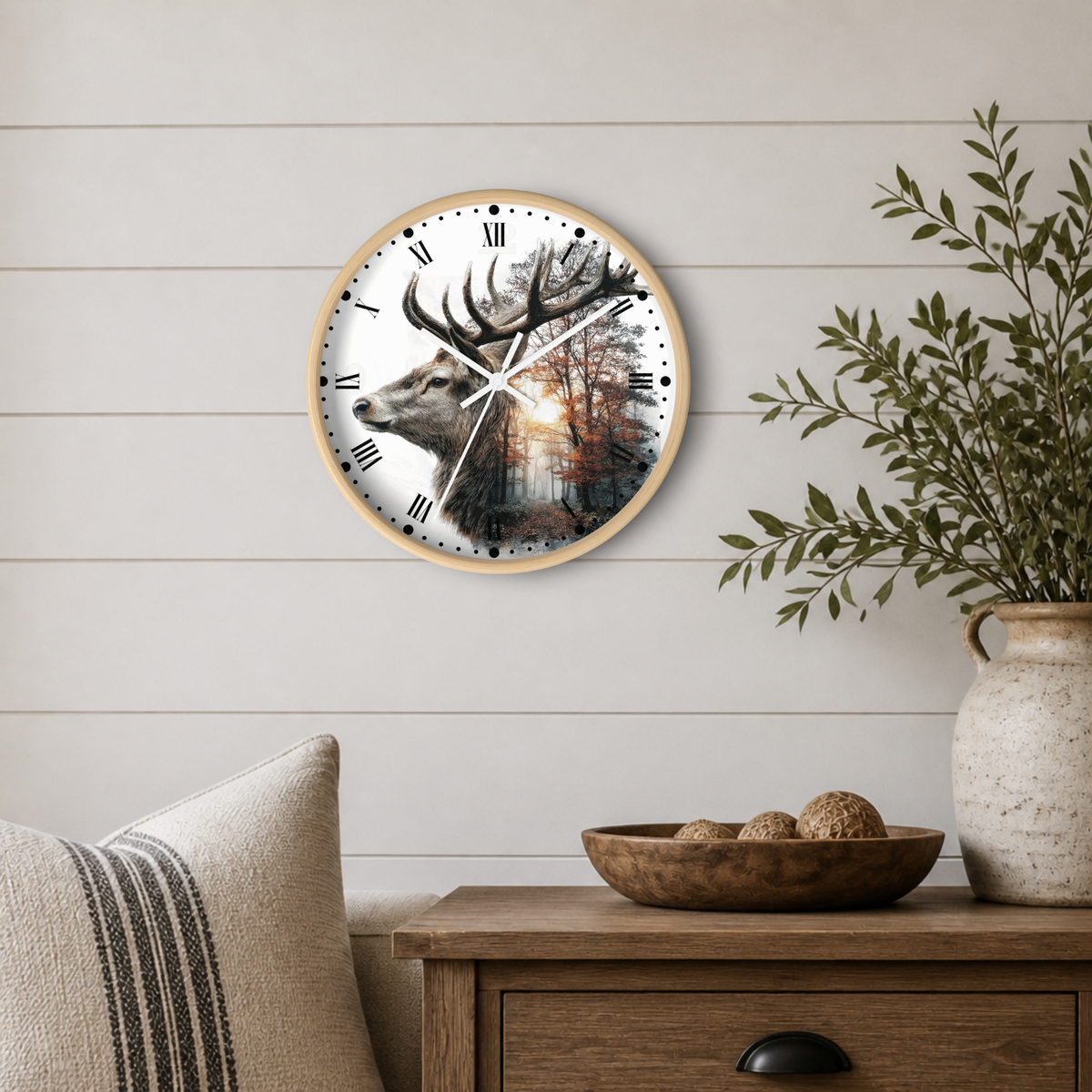 Round wall clock with a light wood frame and a printed deer and forest scene on the face, mounted on a white shiplap wall above a wooden side table with a vase and bowl.