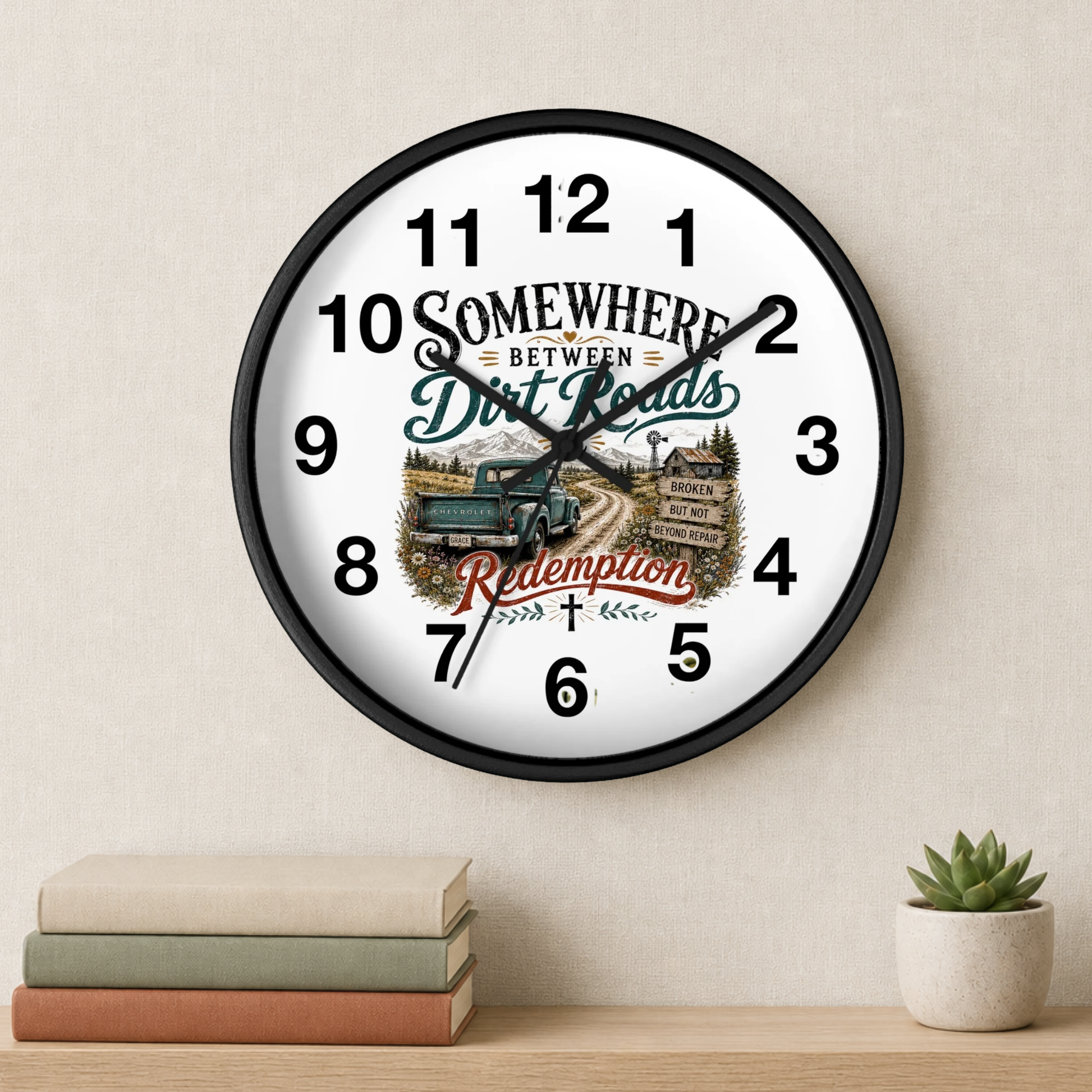 A round wall clock with a black frame and a white face featuring a rustic road scene, a blue pickup truck, and the text 'Somewhere Between Dirt Roads Redemption.' The clock is mounted on a beige wall above a shelf with stacked books and a small potted succulent.