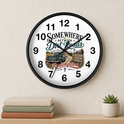 A round wall clock with a black frame and a white face featuring a rustic road scene, a blue pickup truck, and the text 'Somewhere Between Dirt Roads Redemption.' The clock is mounted on a beige wall above a shelf with stacked books and a small potted succulent.