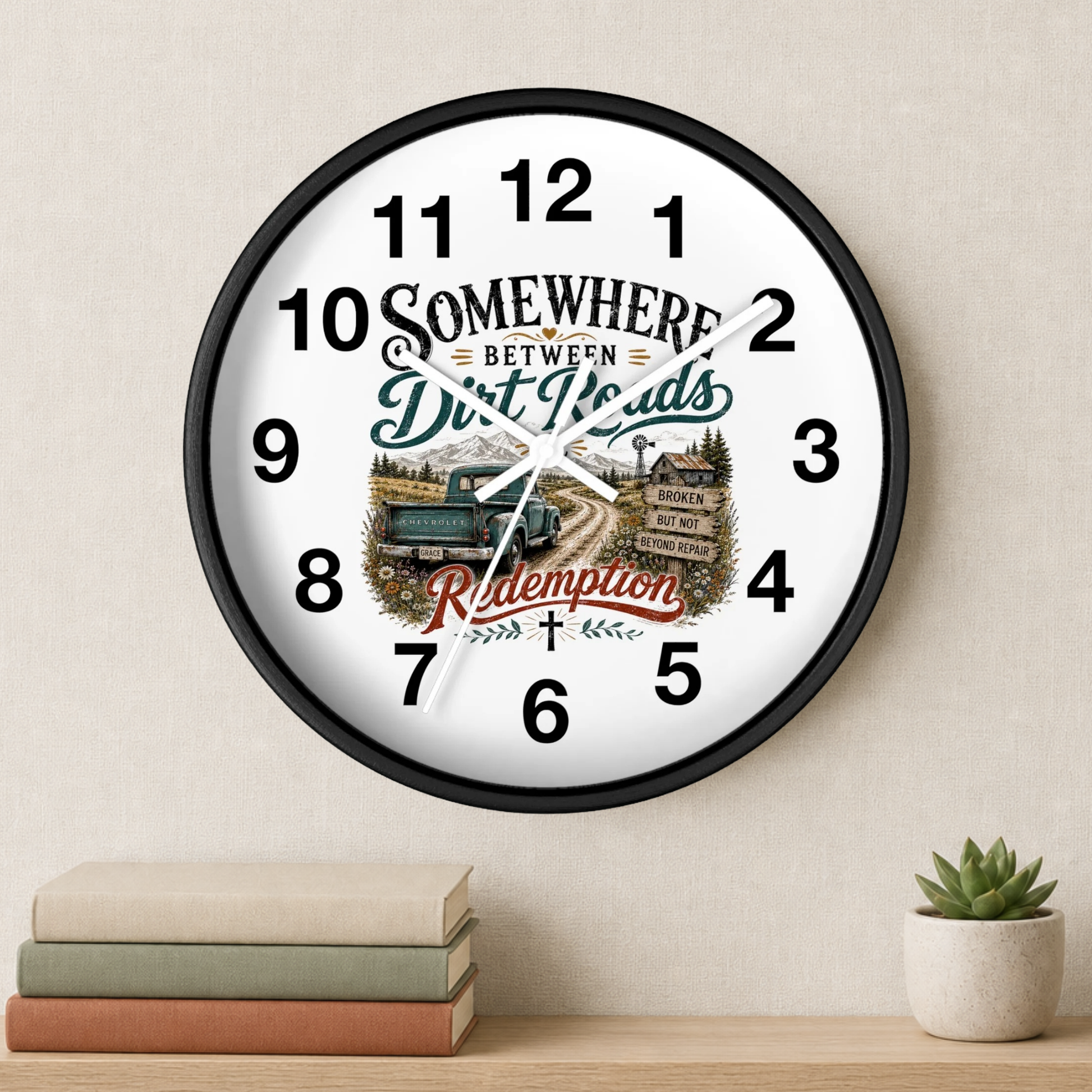 A round wall clock with a black frame and a white face, featuring the phrase 'Somewhere Between Dirt Roads & Redemption' around a vintage truck illustration. The clock is shown hanging on a beige wall above a shelf with books and a small succulent.
