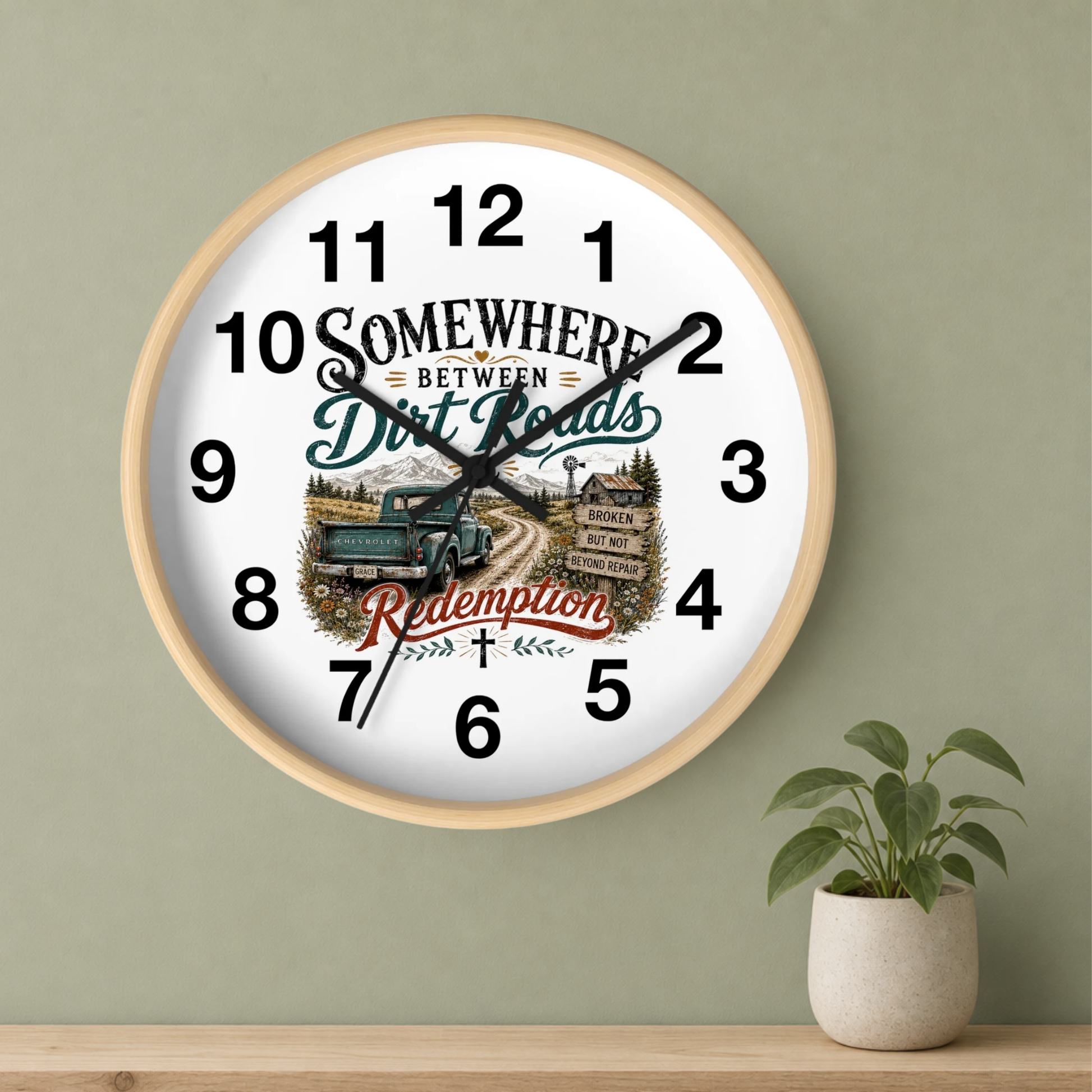 A round wall clock with a light wood frame and a white face, mounted on a sage green wall above a wooden shelf. A small potted green plant sits on the shelf to the right.