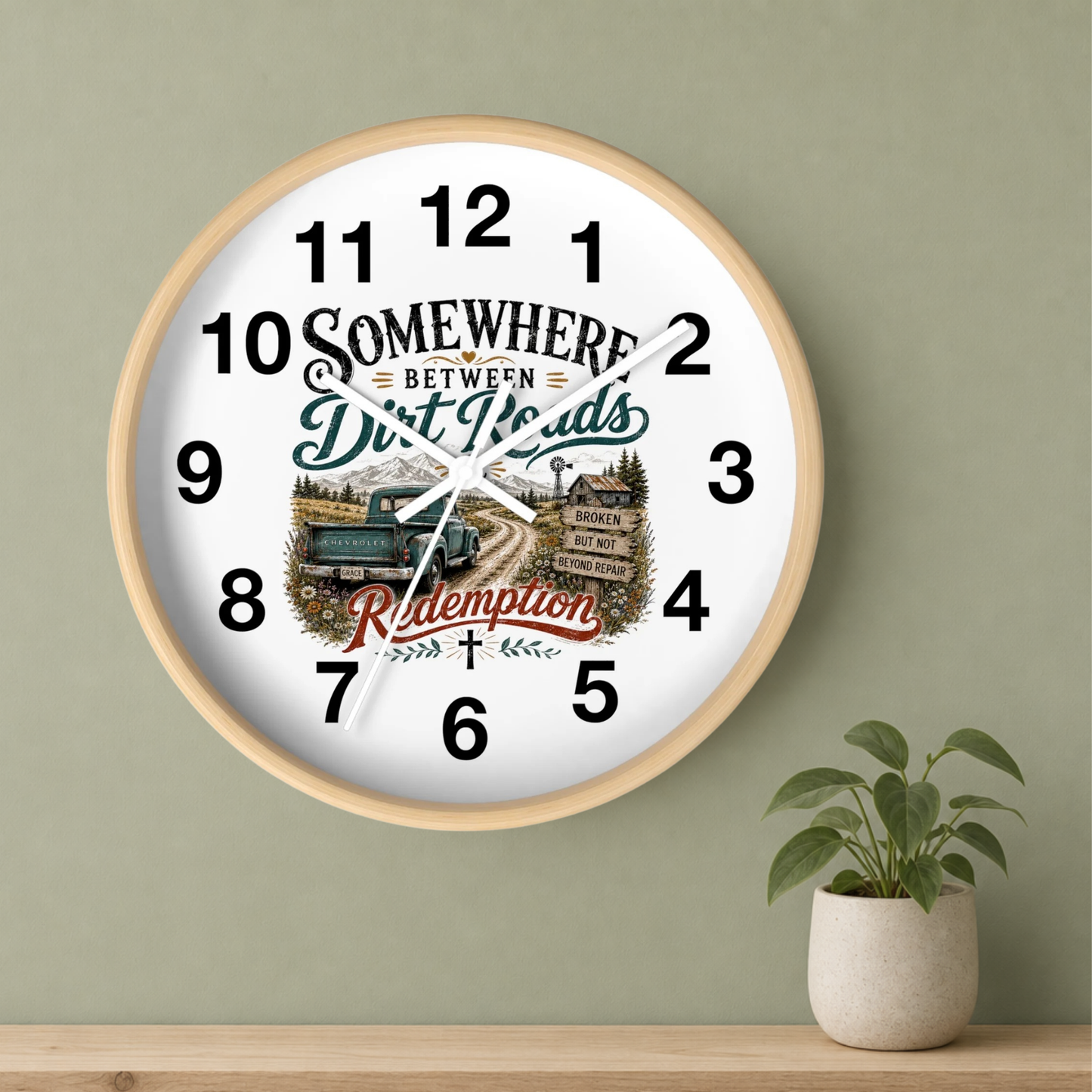 A round wall clock with a light wood frame hangs on a sage green wall above a wooden shelf. A small potted green plant sits on the shelf to the right.