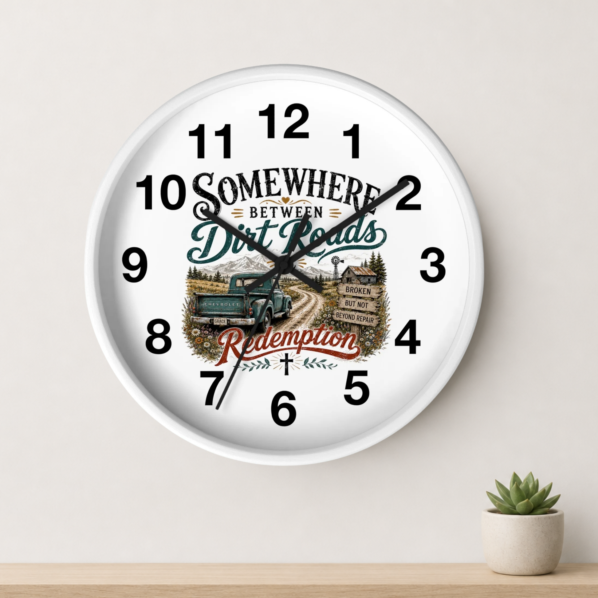 A round white wall clock with black numerals and hands, featuring a rustic western-themed graphic with the words 'Somewhere Between Dirt Roads Redemption' and an illustration of a green truck on a dirt road. The clock is shown hanging on a light beige wall above a wooden shelf with a small potted succulent.