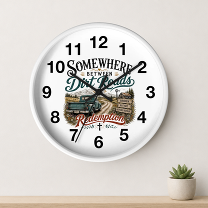 A round white wall clock with black numerals and hands, featuring a rustic western-themed graphic with the words 'Somewhere Between Dirt Roads Redemption' and an illustration of a green truck on a dirt road. The clock is shown hanging on a light beige wall above a wooden shelf with a small potted succulent.