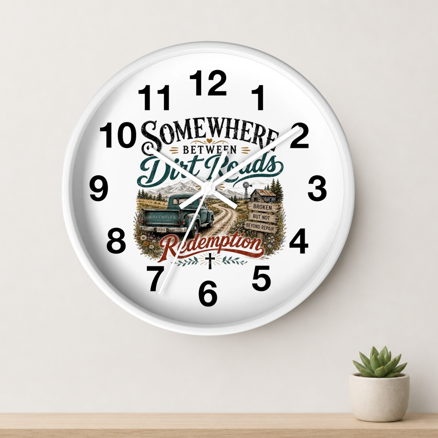 A round white wall clock with black numerals and a rustic western-themed graphic in the center reading 'Somewhere Between Dirt Roads' and 'Ride Again,' shown hanging on a light beige wall above a wooden surface with a small potted succulent.