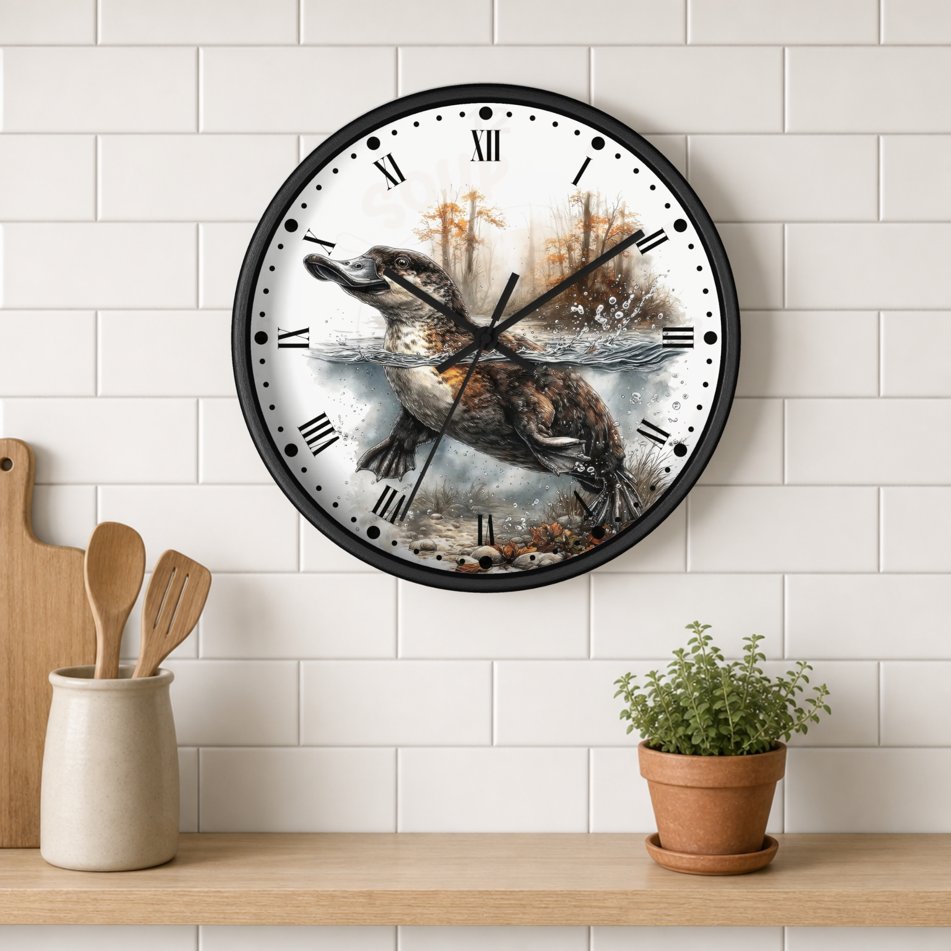 A round wall clock with a black frame and Roman numerals, featuring a detailed illustration of a duckbill platypus hunting by a river. The clock is mounted on a white subway tile wall above a wooden shelf with kitchen utensils and a potted plant.