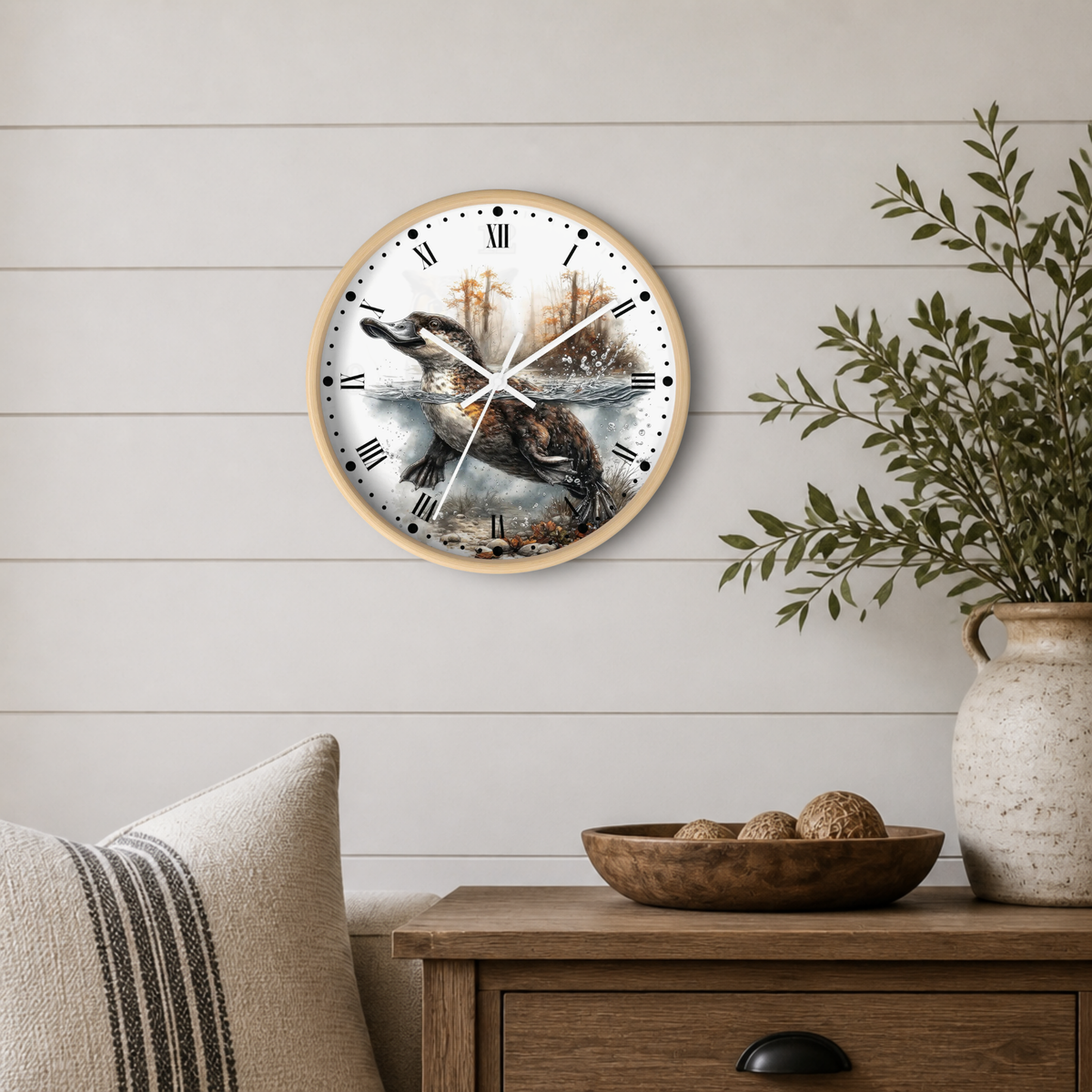 A round wall clock with a natural wood frame hangs on a white shiplap wall above a wooden side table. The clock face features a detailed illustration of a duckbill platypus hunting in shallow water, with Roman numerals and black hands.