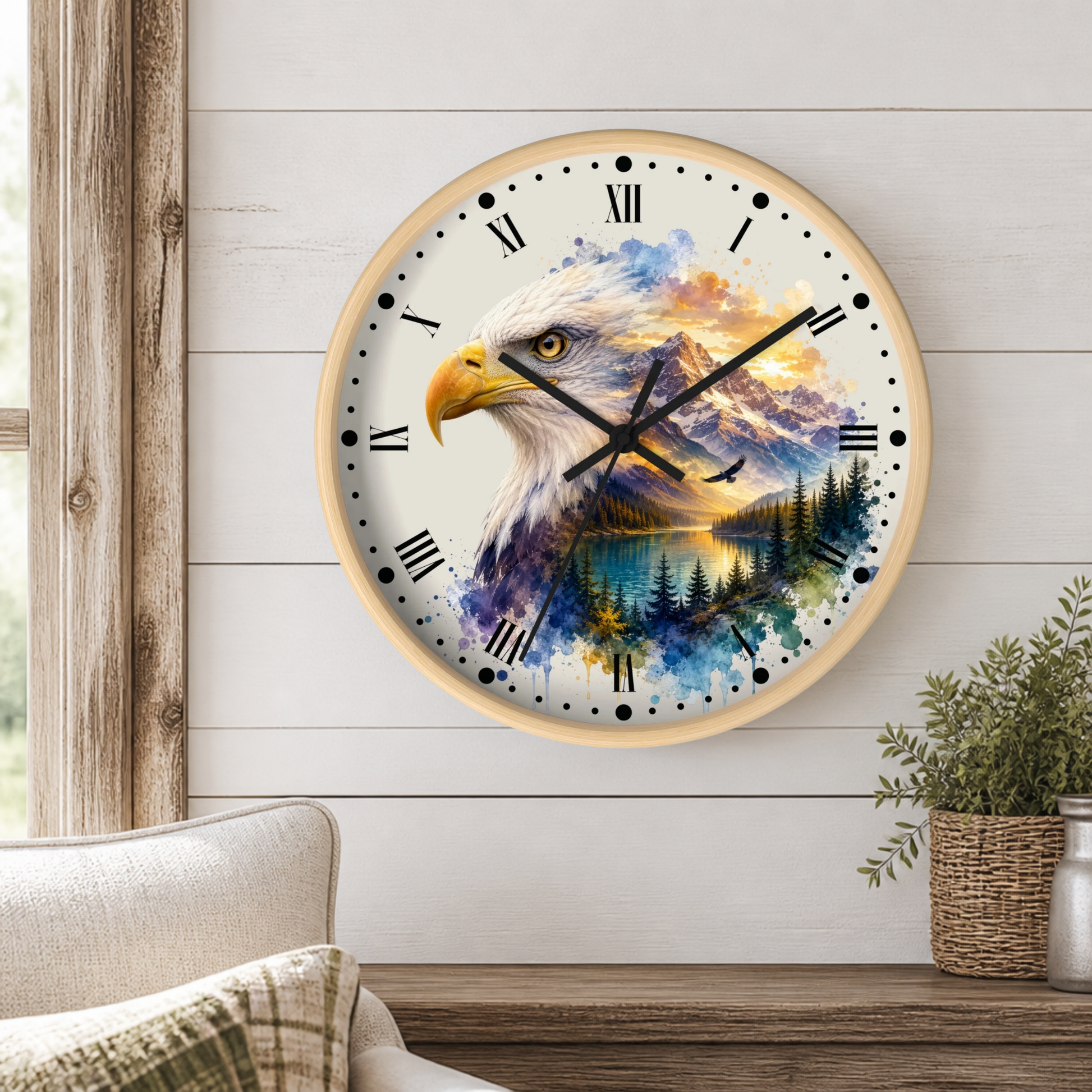 A round wall clock with a light wood frame hangs on a white shiplap wall above a wooden table. The clock face features a detailed watercolor-style design of a bald eagle over a mountain and lake landscape.