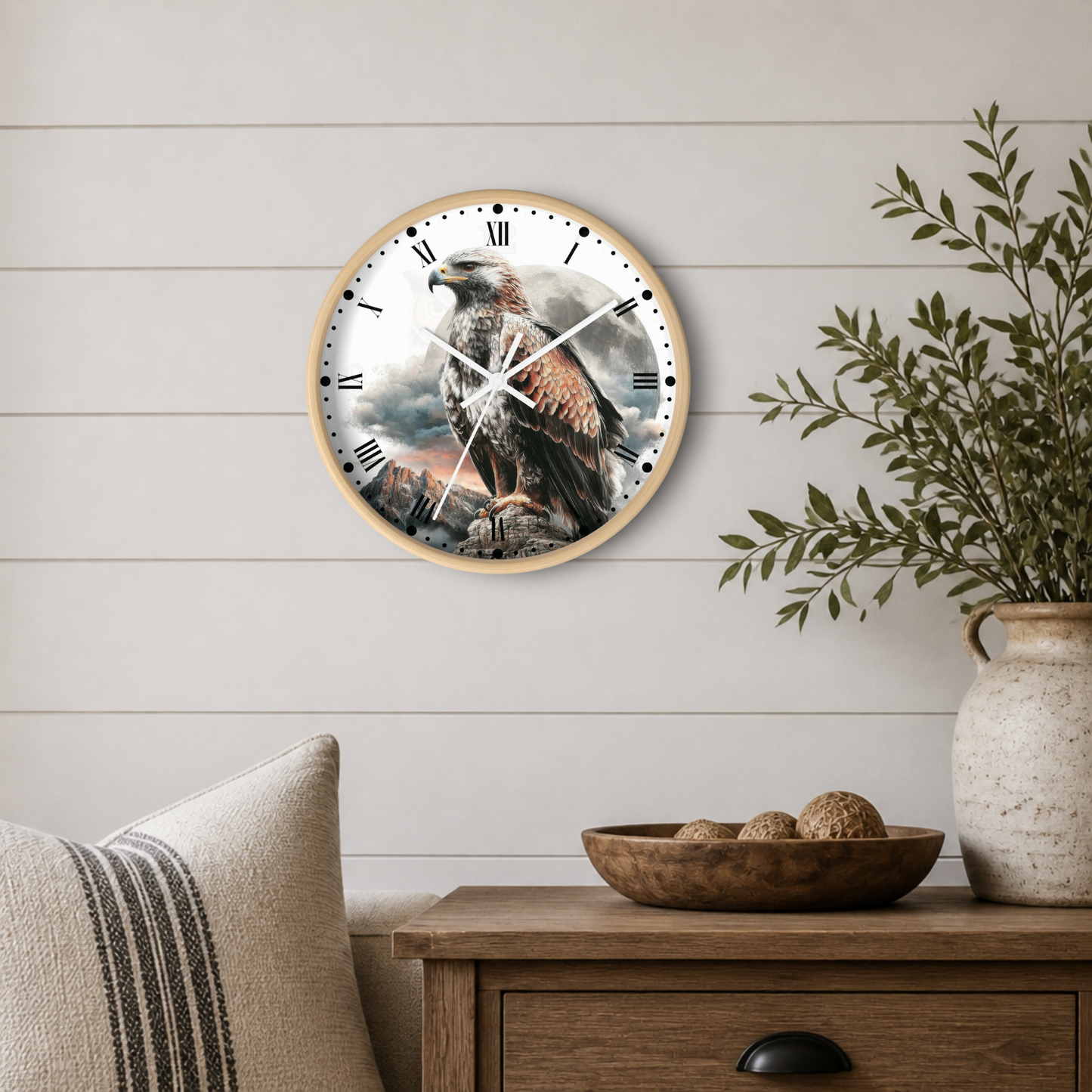 Round wall clock with a natural wood frame featuring a detailed print of a golden eagle perched on a rocky cliff. The clock is mounted on a white shiplap wall above a wooden side table decorated with a bowl, a vase with greenery, and a striped pillow.
