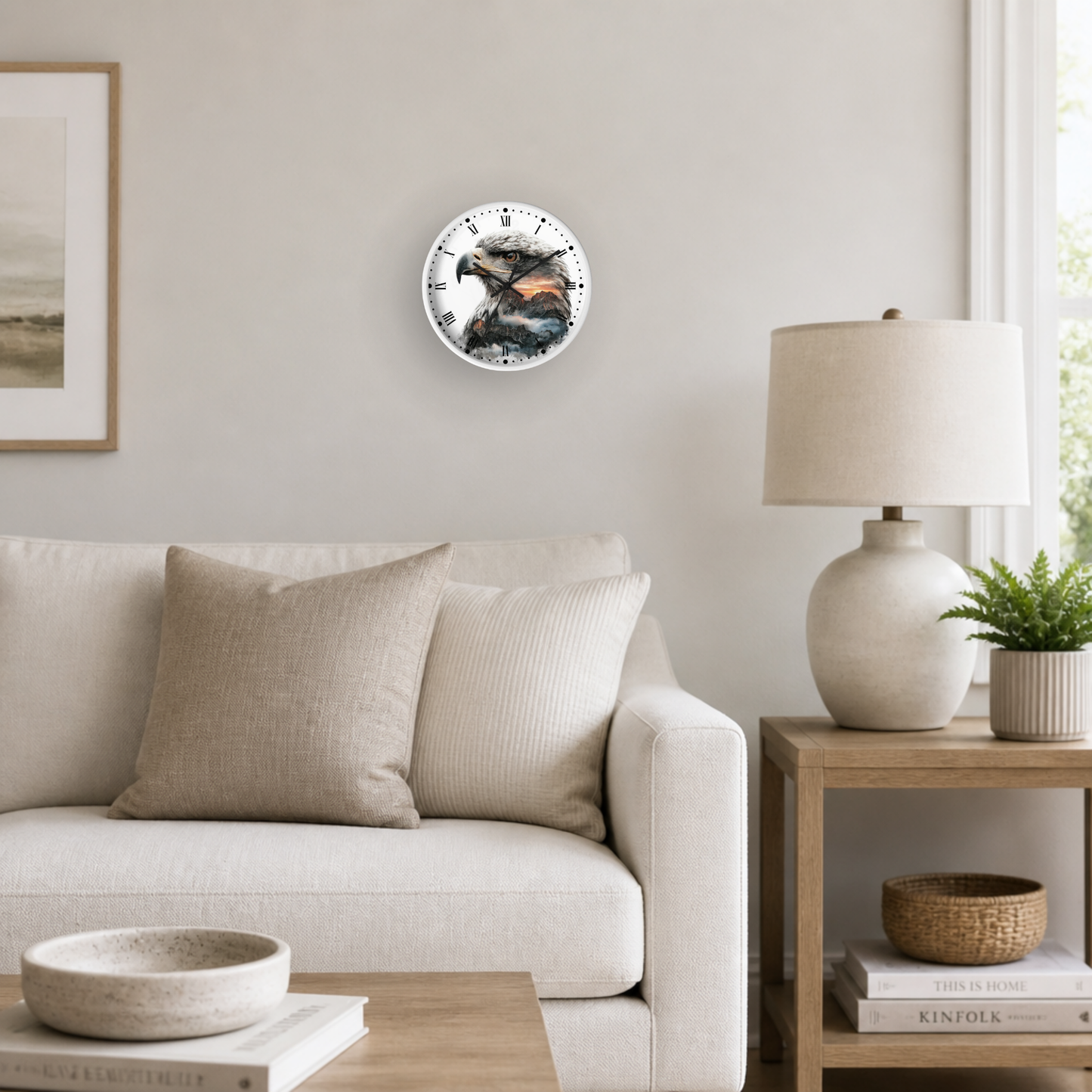 A round wall clock with a colorful mountain and eagle illustration, mounted on a light beige wall in a modern living room setting with a sofa, side table, and lamp.