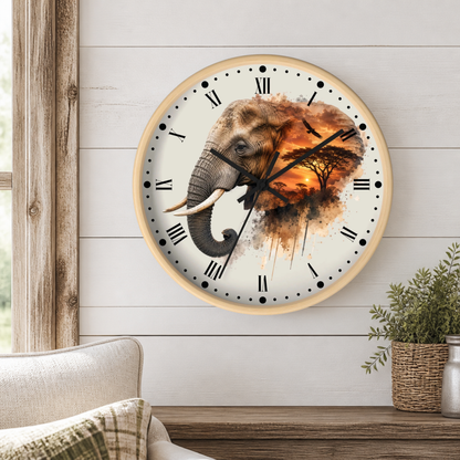 A round wall clock with a light wood frame and Roman numerals, featuring a watercolor-style elephant and savanna sunset design on the face. The clock is mounted on a white shiplap wall above a wooden console table, with a chair and potted plant visible in the room setting.