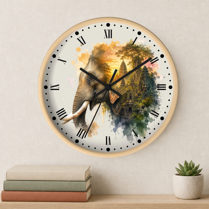 A round wall clock with a light wood frame hangs on a beige wall above a shelf with stacked books and a small potted succulent. The clock face features a watercolor-style elephant merging into a temple scene with gold, green, blue, and pink tones, black Roman numerals, and black hands.