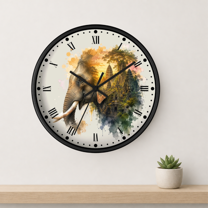 A round wall clock with a black frame and a watercolor-style elephant design on the face, mounted on a light wall above a wooden shelf with a small potted succulent.