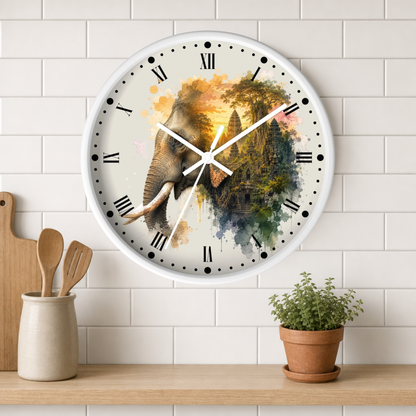 A round wall clock with a white frame and Roman numerals, featuring a watercolor-style elephant and temple design on the face. The clock is mounted on a white subway-tile wall above a wooden shelf with a small potted plant and wooden utensils.