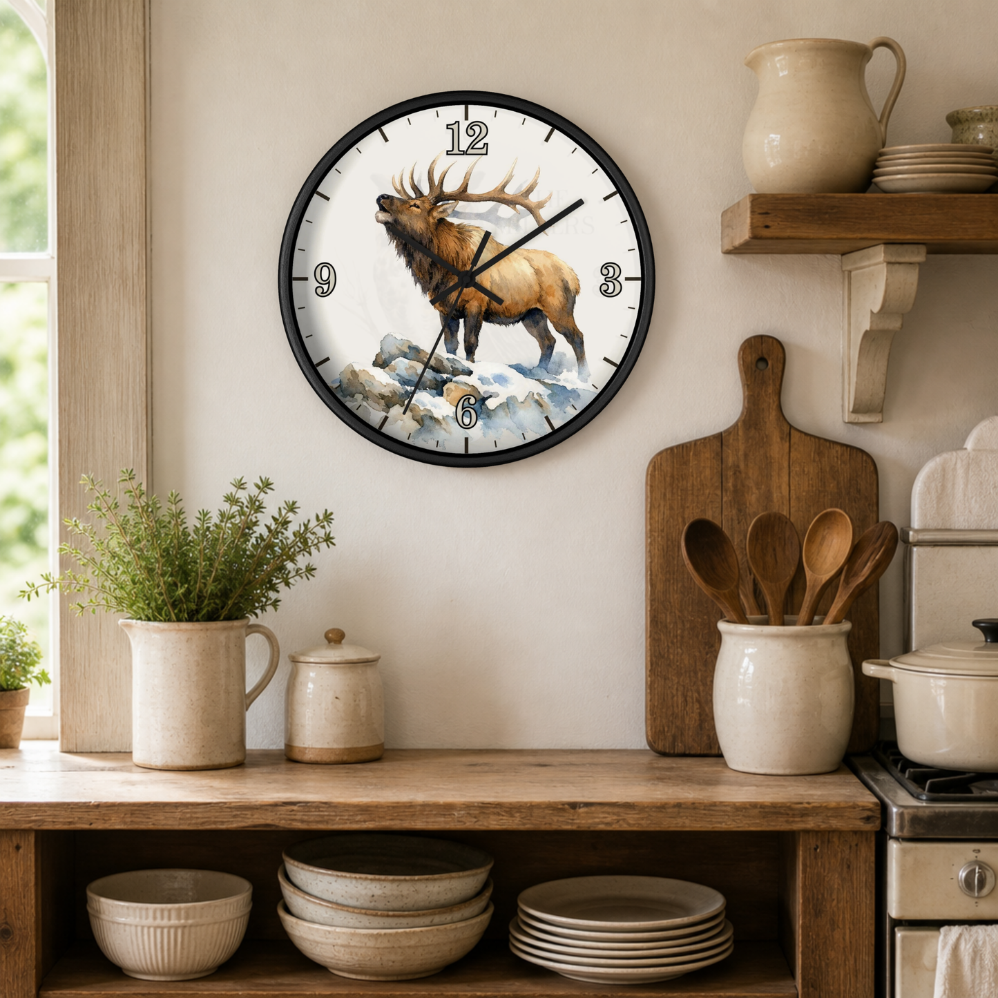 A round wall clock with a black frame and a detailed illustration of a brown elk standing on snowy rocks, mounted on a beige wall in a rustic kitchen setting with wooden shelves, ceramic dishes, and plants.